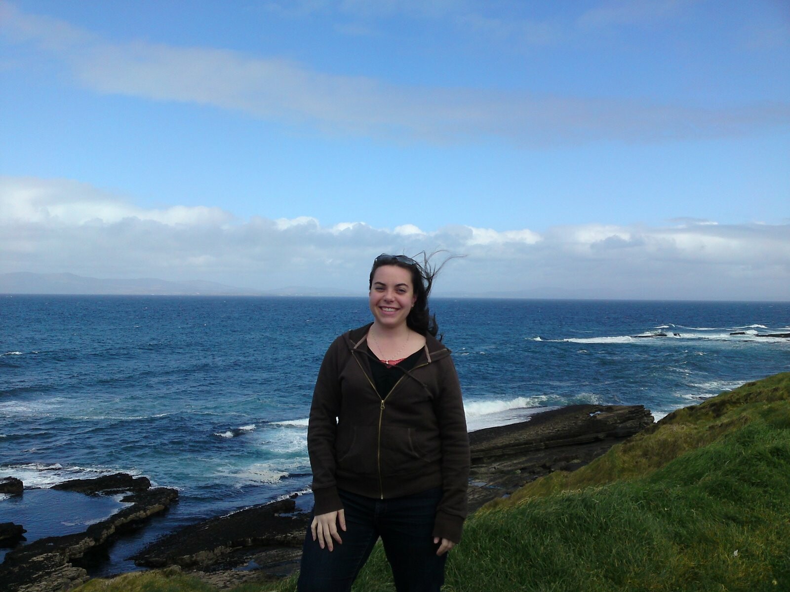 Dacia standing on a seaside cliff, hair blowing in the wind, with the ocean behind her